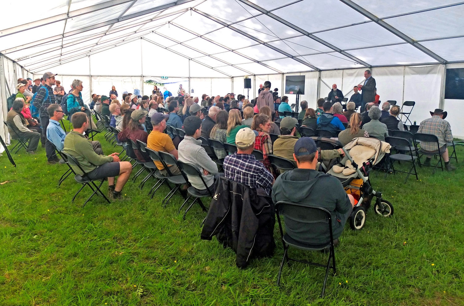 Seated crowd in marque tent listening to speaker. 