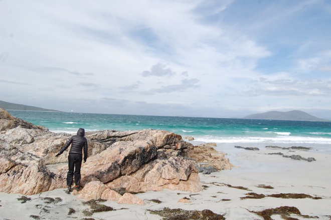 Black-clad figure on rocks on beach with blue sea and sky