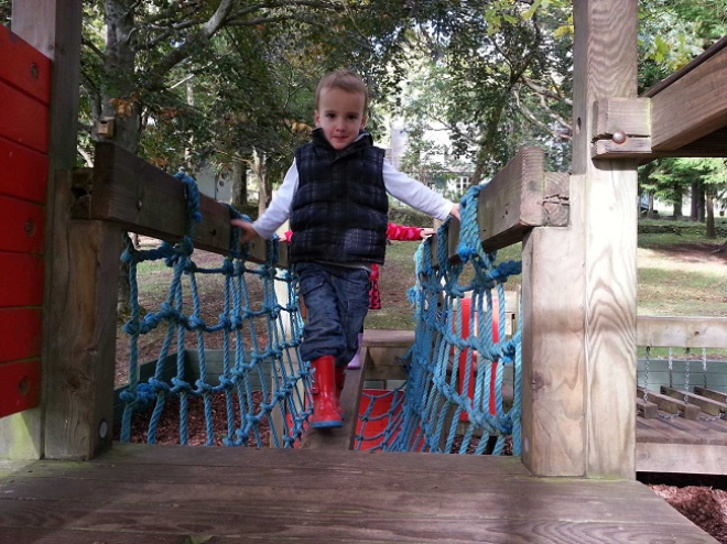 young boy in red wellies balances on wooden play structure