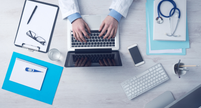 Doctor sitting at table with laptop, papers, phone etc